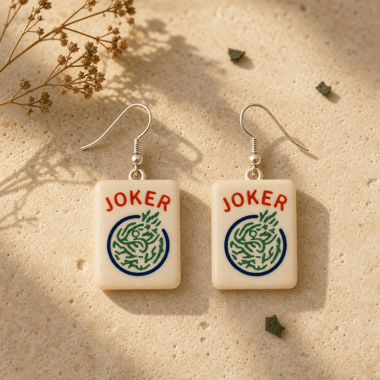 Mahjong Joker Tile Earrings laid on warm sandstone with dried florals, showing the crisp red JOKER lettering and green dragon medallion